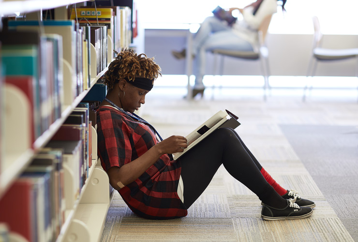 Student sitting on floor in library reading book.